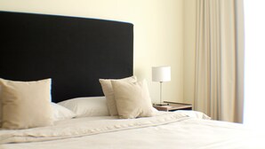 A neatly made bed with white linens and two beige pillows, a bedside table with a lamp, and a window with curtains.