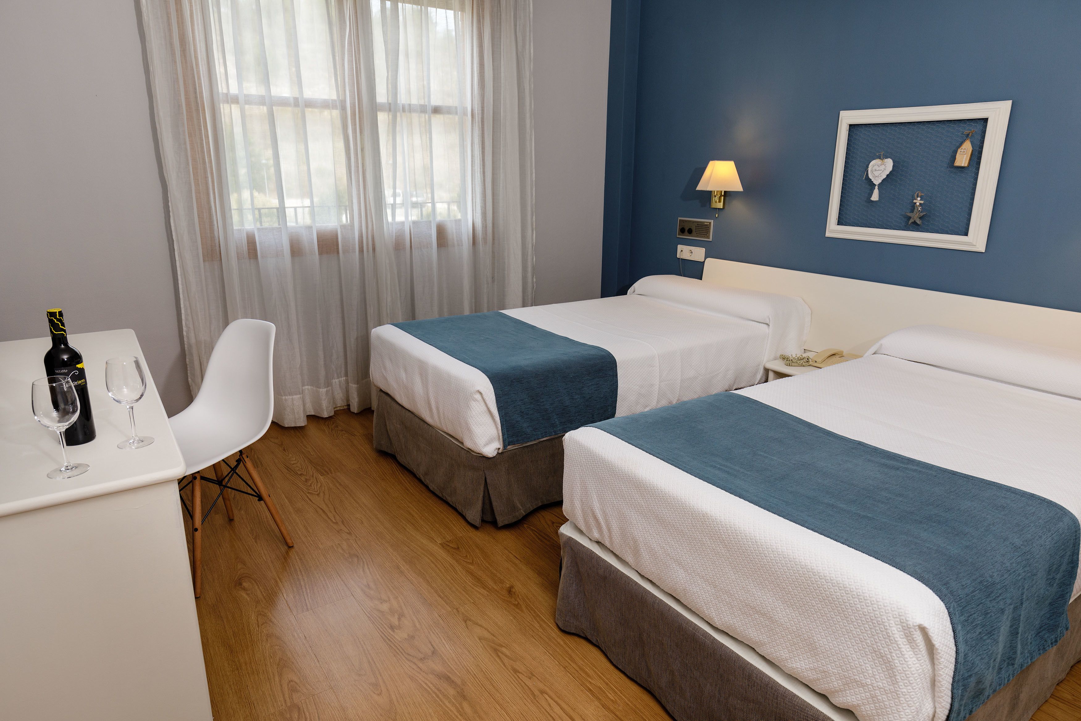 A hotel room with two beds, a desk with a bottle and glasses, a chair, and a wall with a framed picture.
