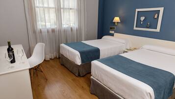 A hotel room with two beds, a desk with a bottle and glasses, a chair, and a wall with a framed picture.