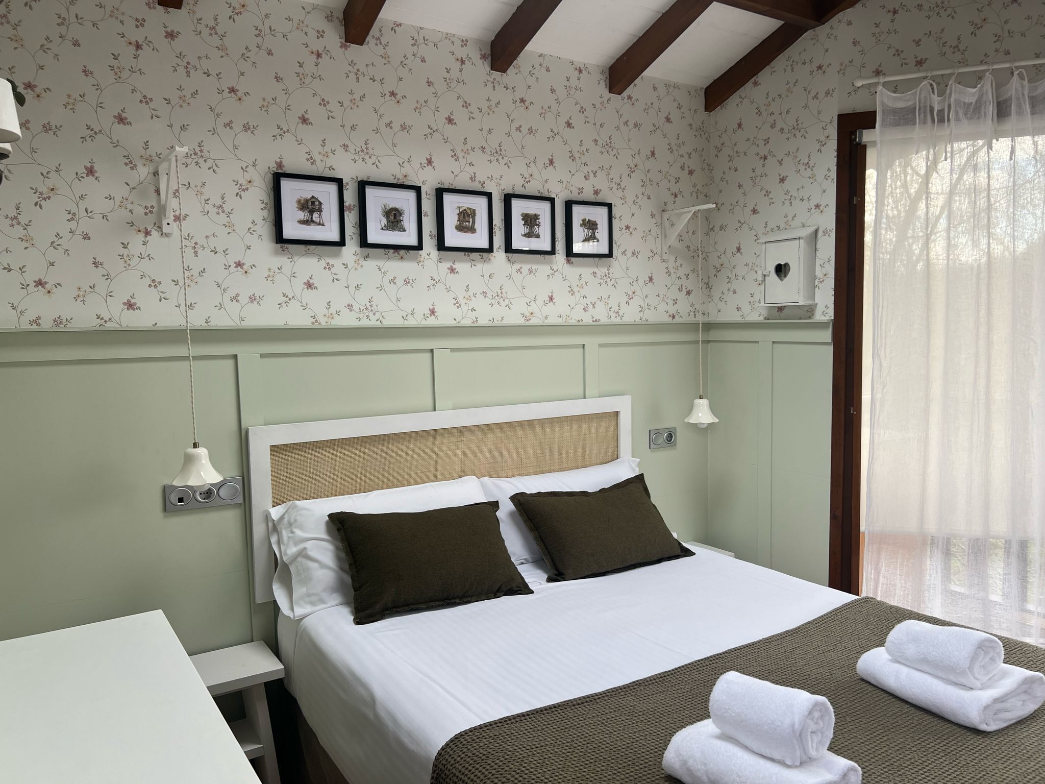Una cama bien hecha con ropa de cama blanca y marrón, un cabecero con panel tejido, y una pared con papel tapiz floral y cuadros enmarcados.