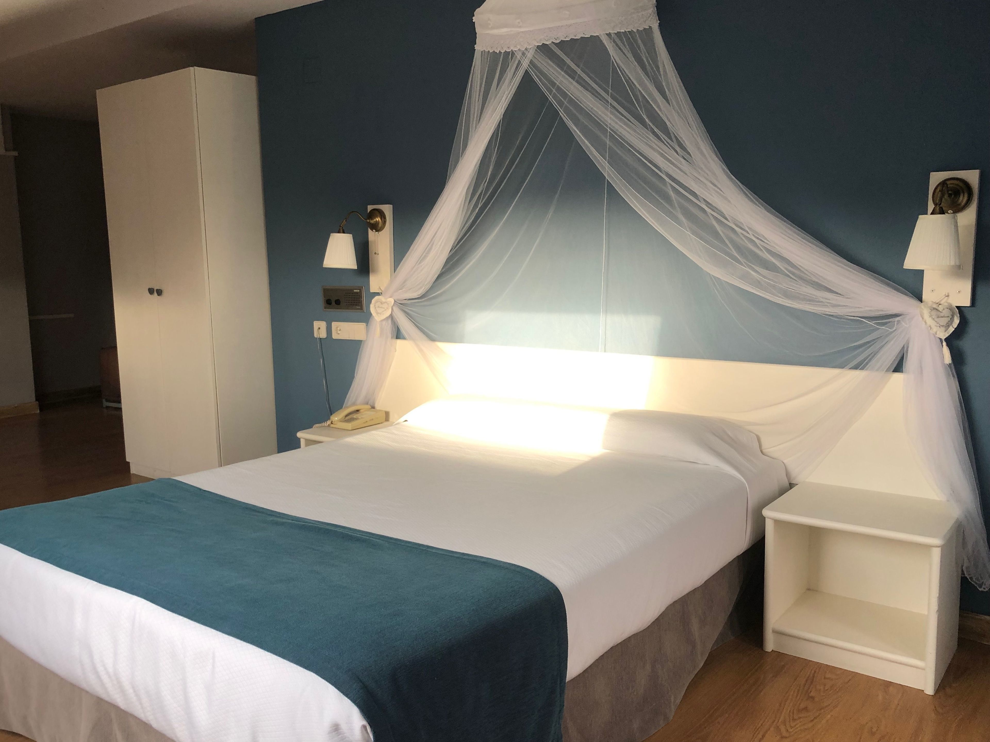 Un lit à baldaquin blanc, une table de chevet, une commode, une armoire et une lampe fixée au mur.