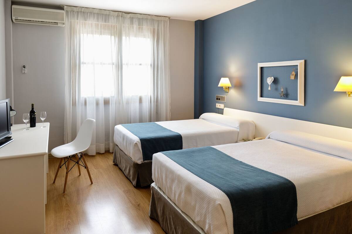 Habitación de hotel con dos camas, un escritorio, una silla y una ventana con cortinas translúcidas.