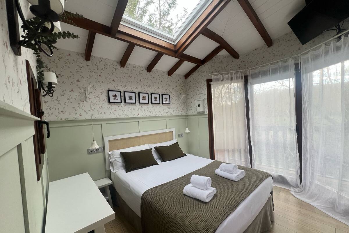 Un dormitorio con cama, mesitas de noche, escritorio, ventana con cortinas traslúcidas y tragaluz.