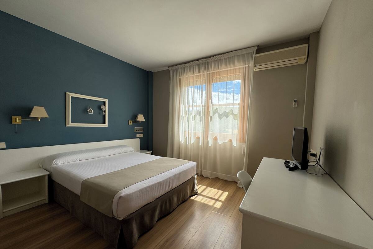 Habitación de hotel con cama, mesitas de noche, escritorio con computadora y ventana con cortinas.