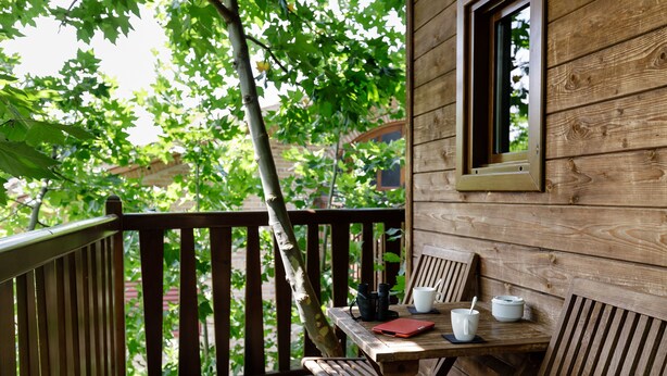 Eine Holzterrasse mit Tisch und Stühlen, ein Fenster und im Hintergrund Bäume sowie ein Gebäude.
