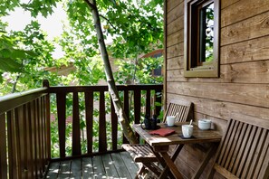 A wooden deck with a table and chairs, a window, and a view of trees and a building in the background. - Hotel Jakue (Puente La Reina)
