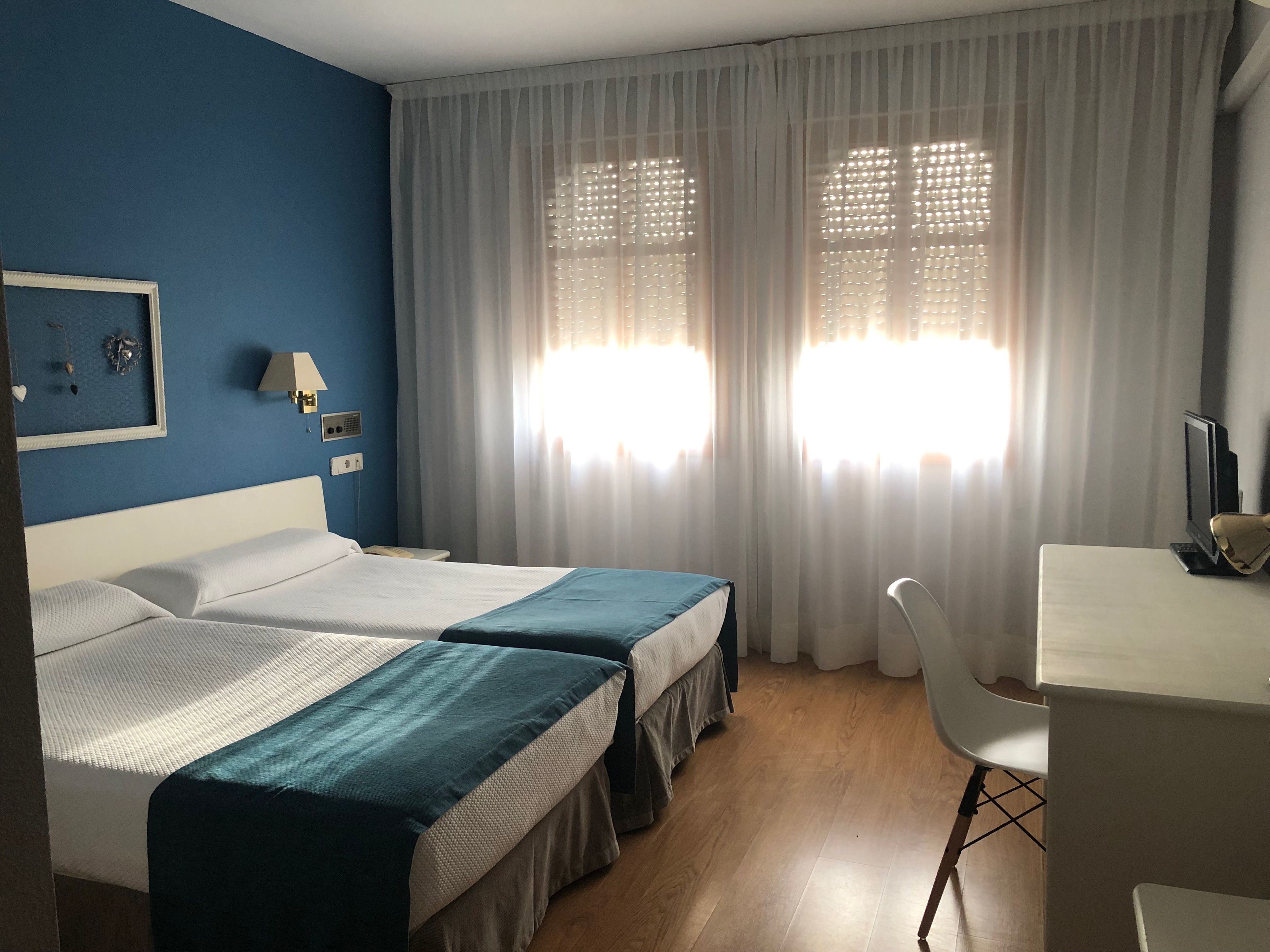 Ett hotellrum med två sängar, ett skrivbord, en stol och ett fönster med tunna, genomskinliga gardiner.