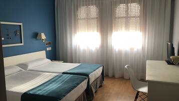 Ett hotellrum med två sängar, ett skrivbord, en stol och ett fönster med tunna, genomskinliga gardiner.