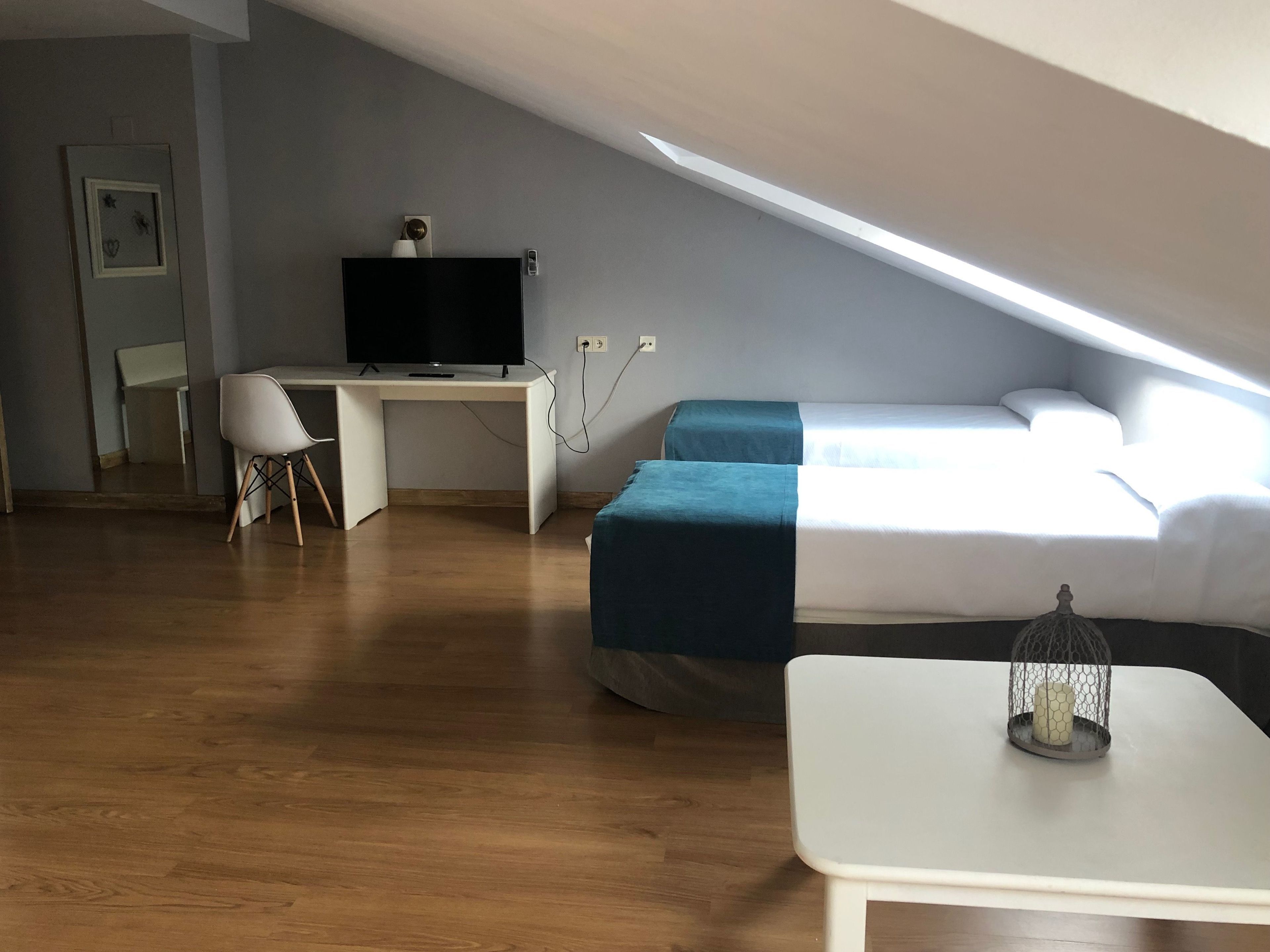 Habitació moderna i compacte amb sostre inclinat, escriptori blanc amb televisió de pantalla plana, llit amb llençol blau i taula petita amb un objecte decoratiu.