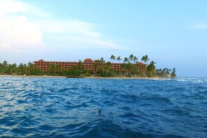 Un hotel frente a la playa con varios pisos, palmeras y vista al océano.