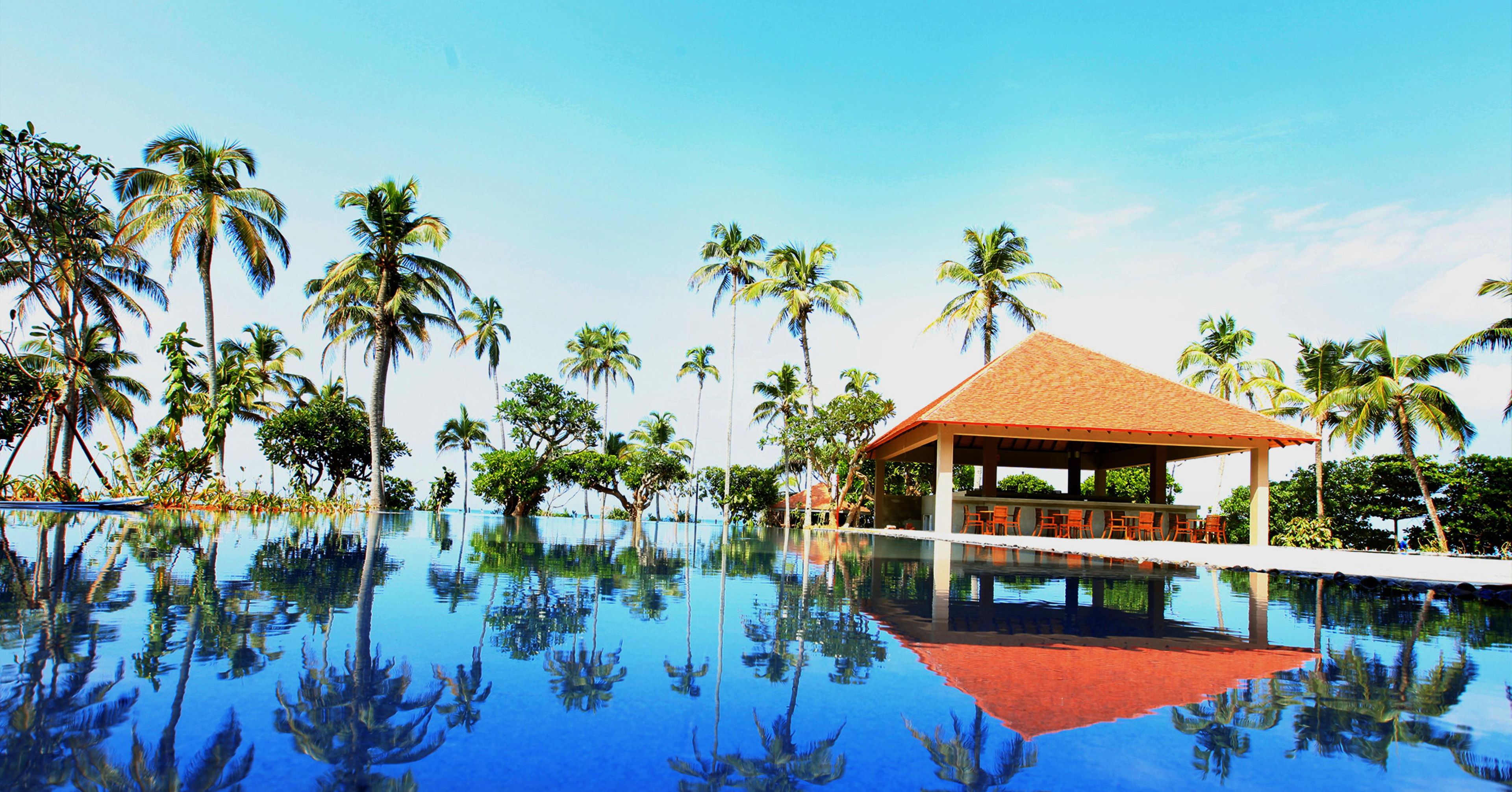 A clear blue pool with reflections of palm trees, surrounded by a covered seating area with orange chairs.