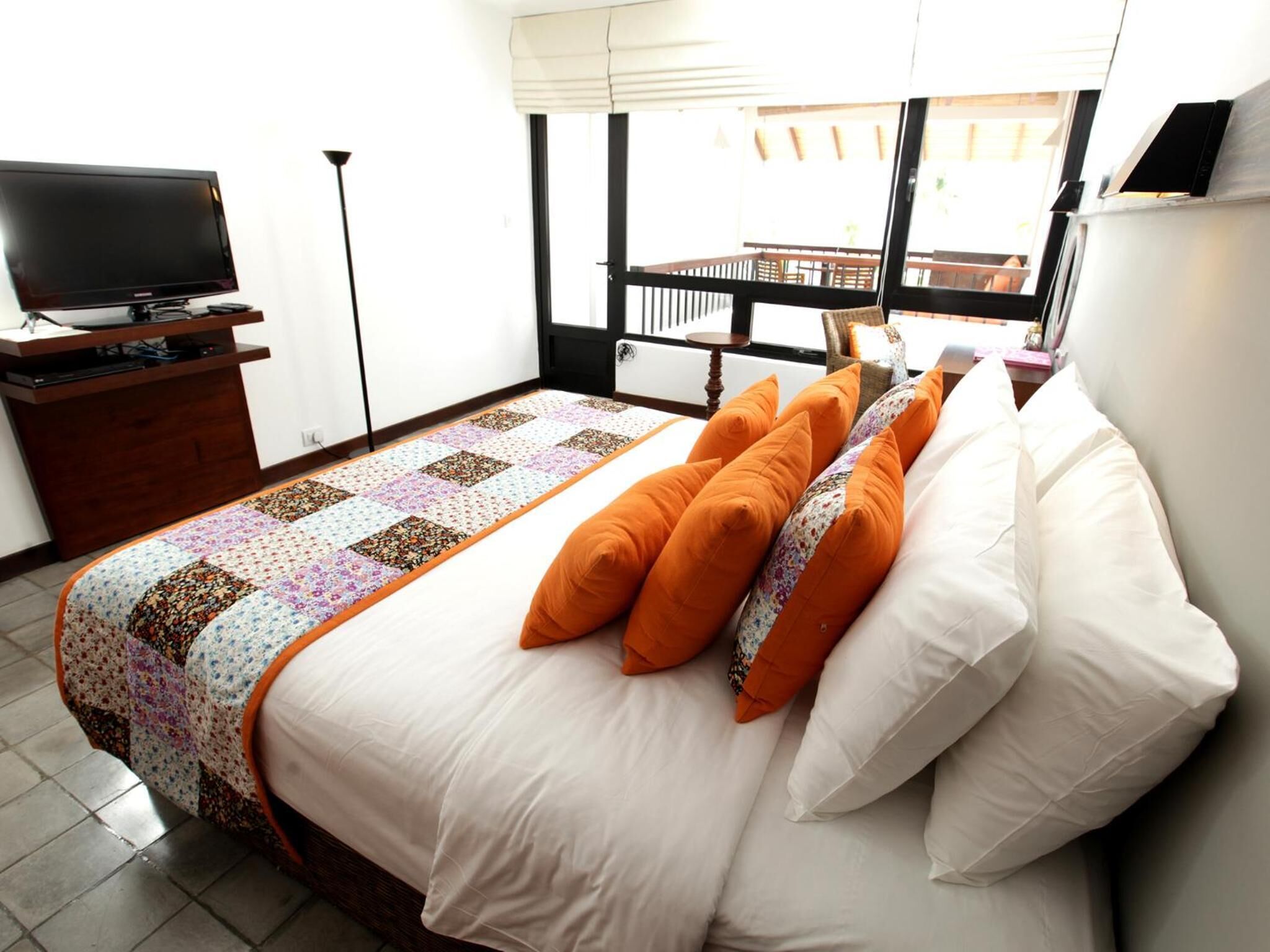 Una cama bien hecha con almohadas blancas y anaranjadas, una colcha y un televisor sobre un soporte de madera en una habitación luminosa con vista a un balcón.
