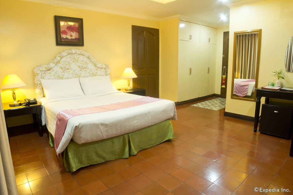 Habitación de hotel con una cama, dos mesitas de noche con lámparas, un tocador, un escritorio con una silla, un armario y un espejo.