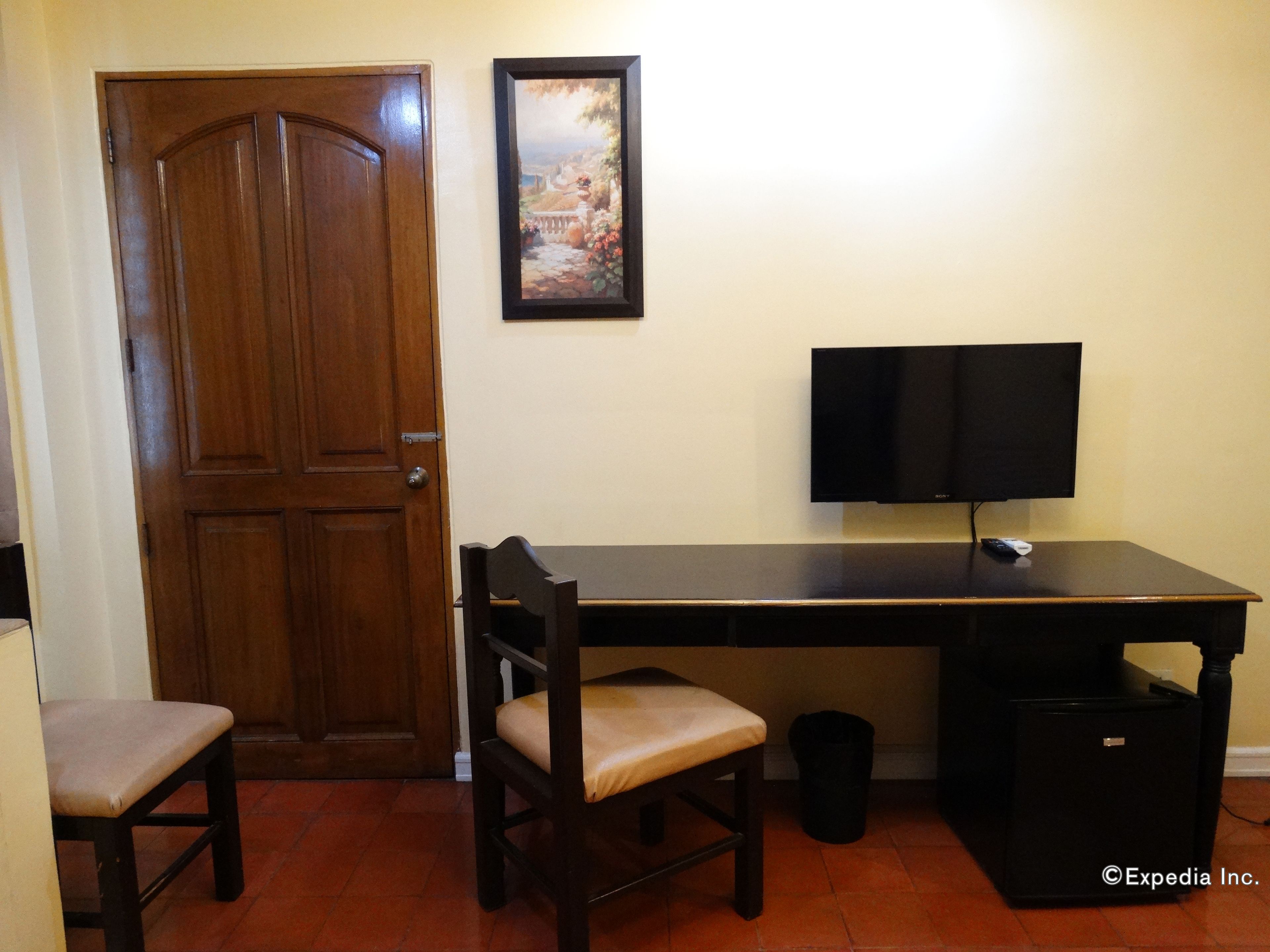 A room with a wooden door, a framed picture on the wall, a desk with a flat-screen TV, a chair, and a trash bin.