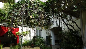 A courtyard with potted plants, a red curtain, and a white building with arches. - White Knight Hotel Intramuros (Manila)