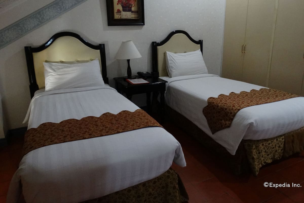 Una habitación de hotel con dos camas, una mesita de noche con lámpara, un cuadro enmarcado en la pared y un armario.