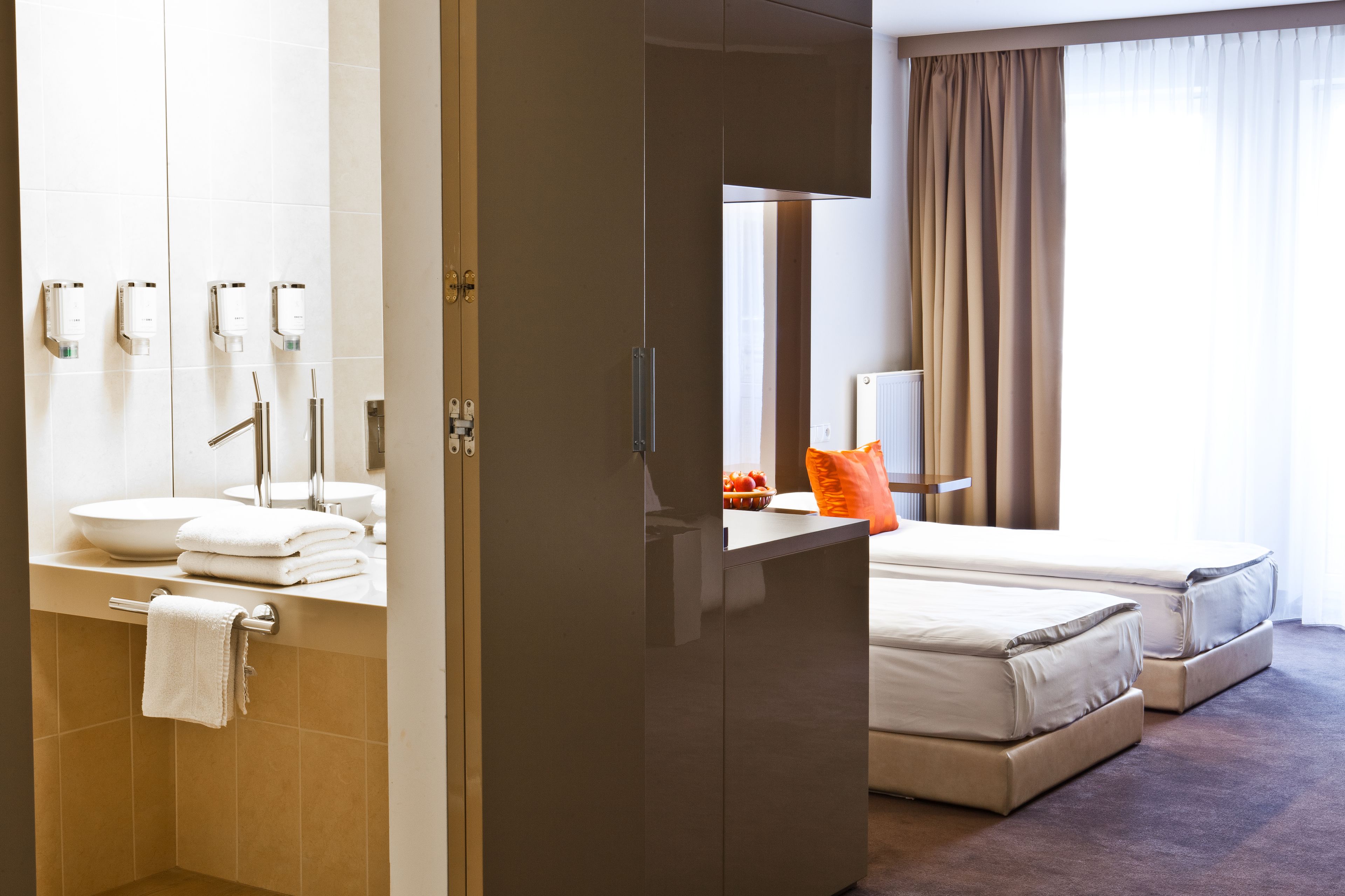 A hotel room with a bathroom featuring a sink, mirror, and towel rack. The room has a bed with two pillows and a nightstand with a lamp.