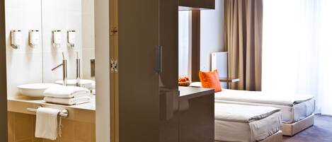 A hotel room with a bathroom featuring a sink, mirror, and towel rack. The room has a bed with two pillows and a nightstand with a lamp.