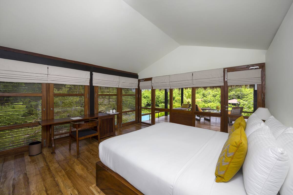 A bedroom with a large bed, wooden desk, and large windows with blinds.