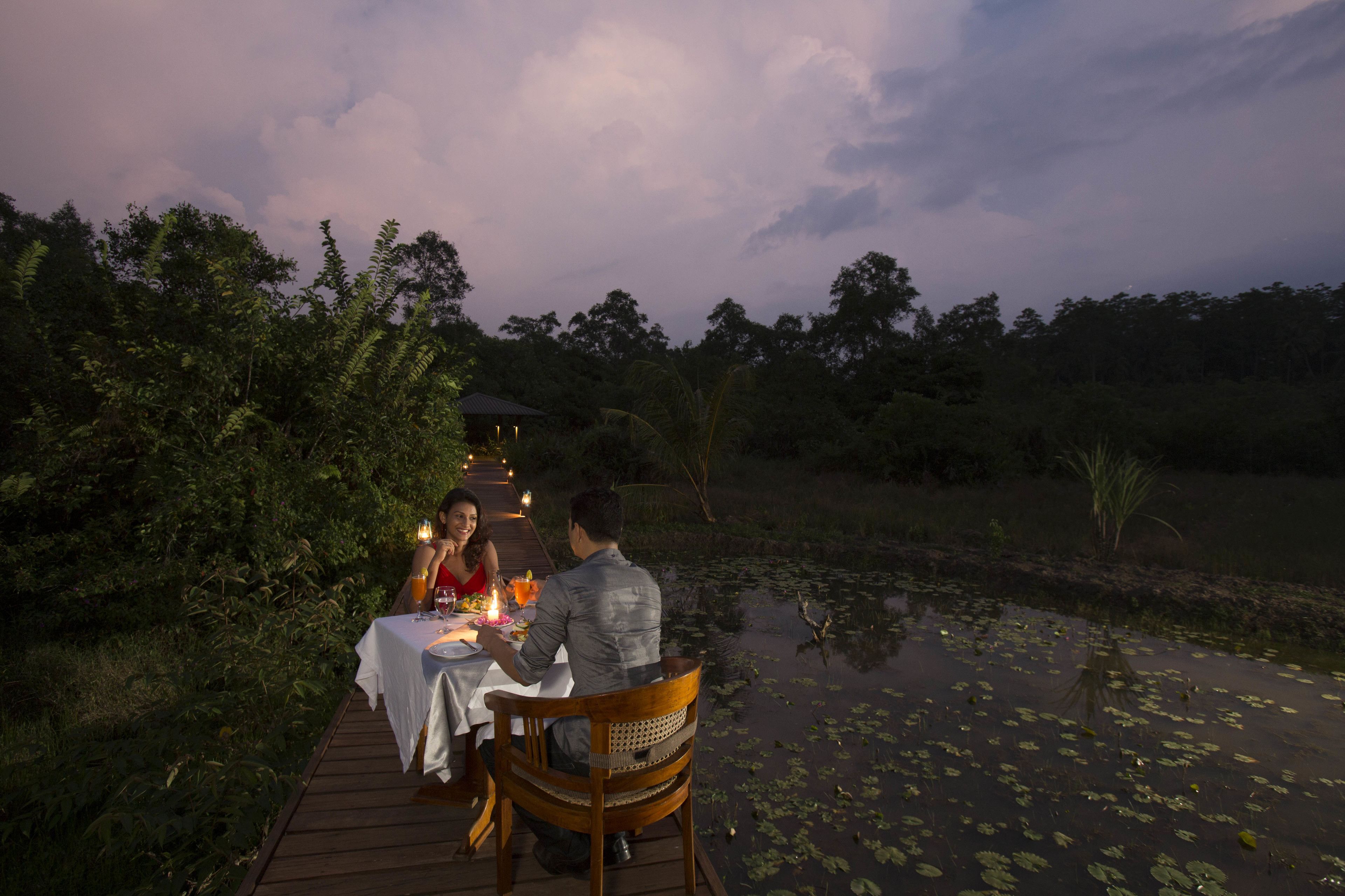 A couple dining at a wooden table by a pond with lily pads, surrounded by greenery.