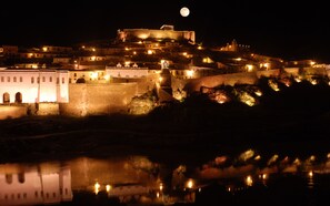 A historic town with a prominent castle on a hill, illuminated by street lights and reflecting in a body of water.