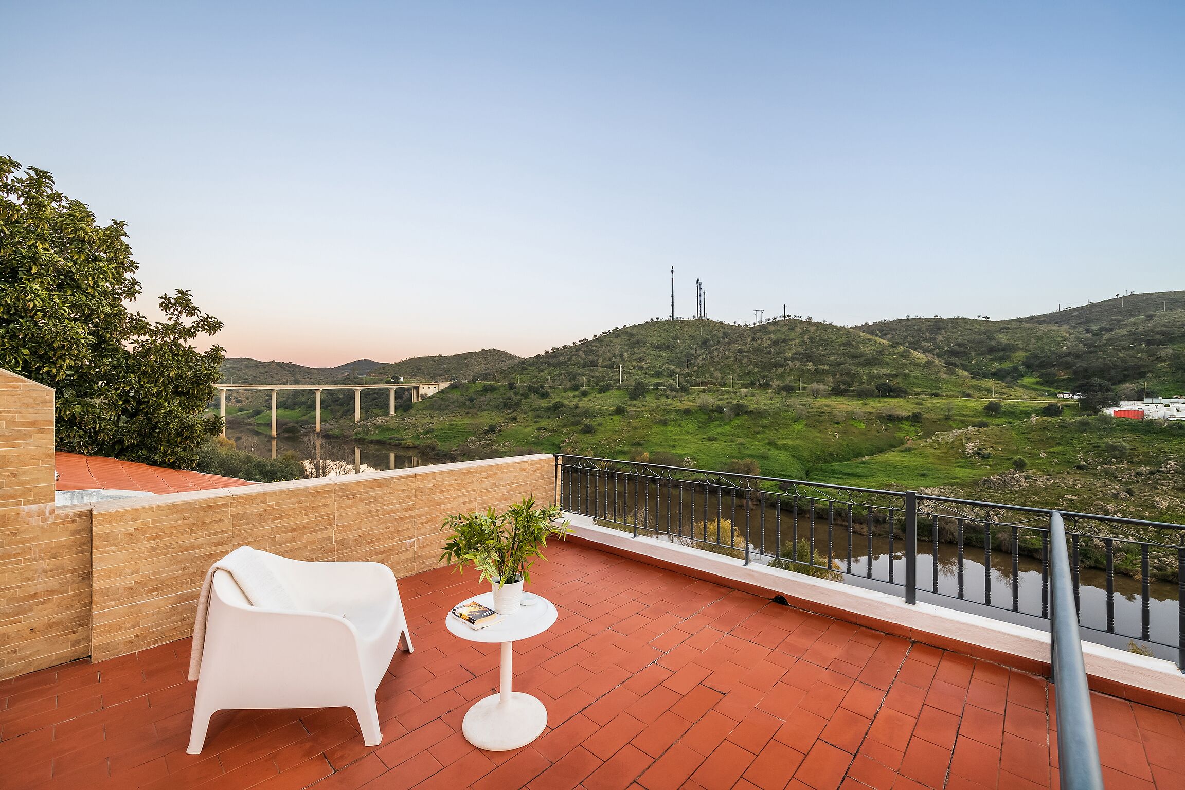 A balcony with a white chair and a small table, overlooking a river and a bridge in a hilly landscape.