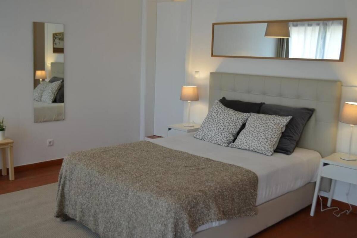 A bedroom with a large bed, two bedside tables with lamps, a mirror above the bed, and a small side table with a plant.