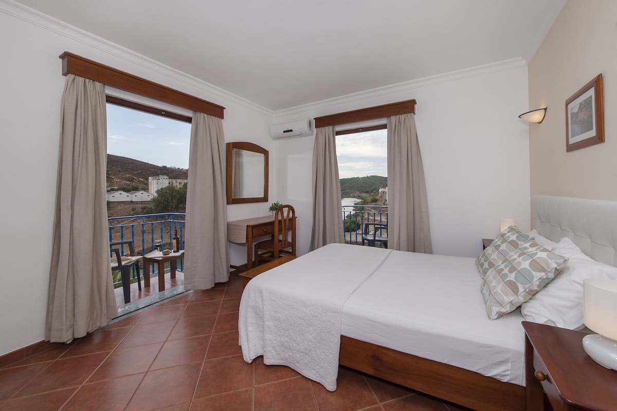 A hotel room with a bed, a desk, and a balcony with a view of the city.