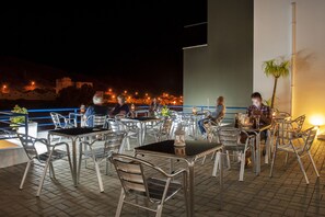 An outdoor dining area with metal chairs and tables, a view of the cityscape in the background, and a few people dining.
