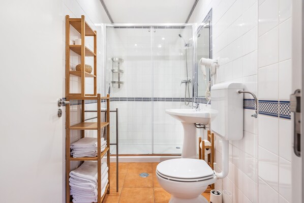 Un baño pequeño con ducha de vidrio, estante de madera con toallas, lavamanos pedestal y un inodoro.