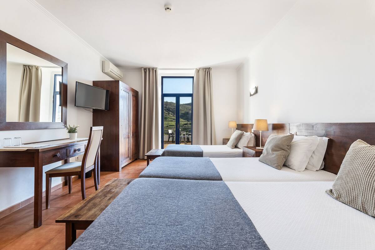 A hotel room with two beds, a desk, a TV, and a large window with a view of greenery.
