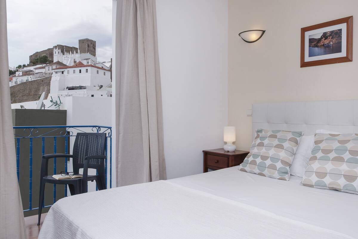 A hotel room with a bed, a chair, a nightstand, a lamp, a picture on the wall, and a view of a castle.