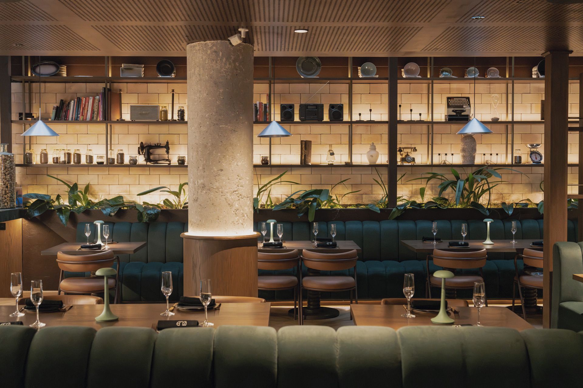 L’intérieur d’un restaurant présente un mur en briques, des étagères remplies d’ustensiles de cuisine et des sièges recouverts d’un tissu vert.