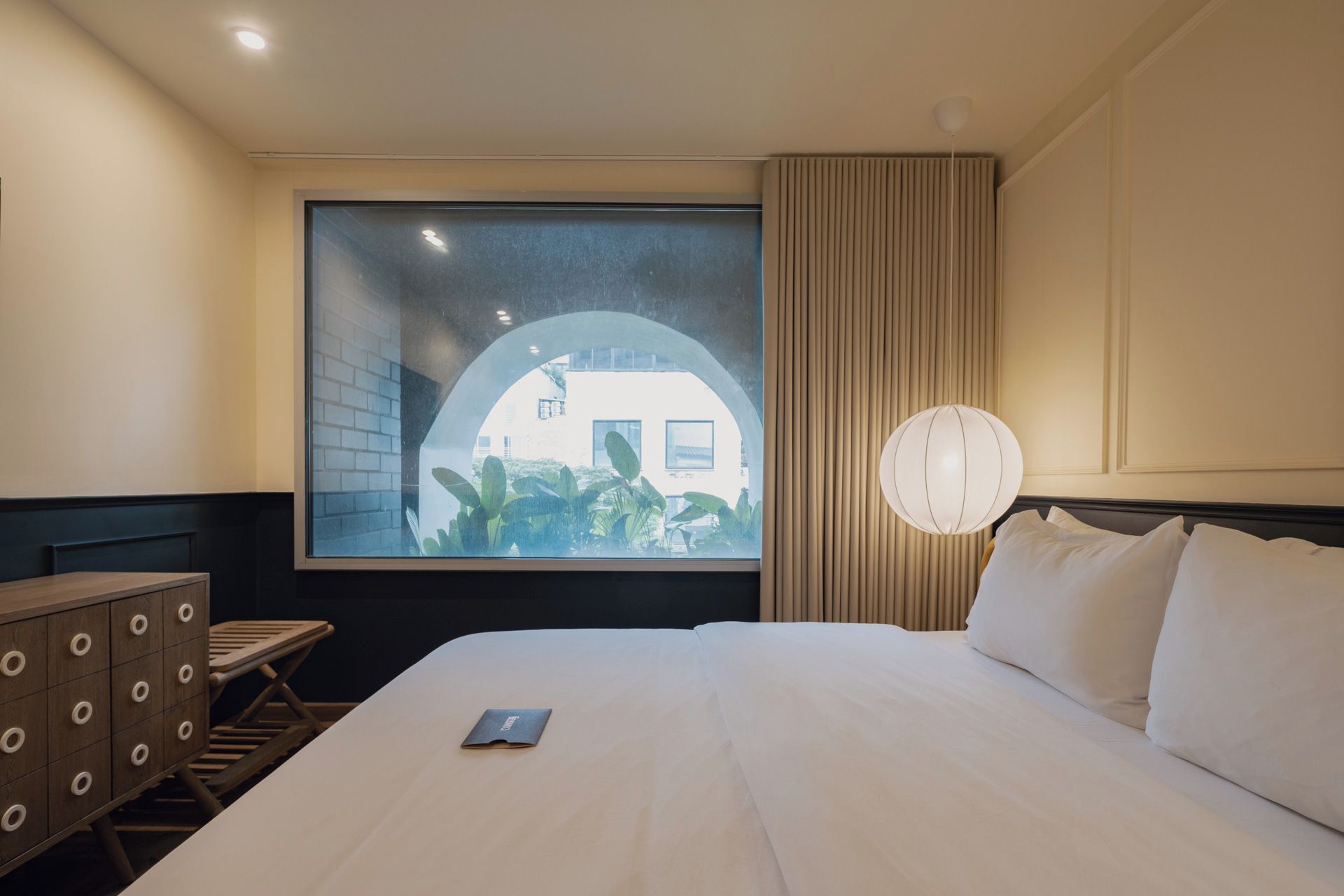 A hotel room with a bed, a desk, a chair, a window with a view of a building, and a lamp.