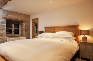 Un dormitorio con pared de piedra, cabecero de madera, cama con ropa blanca, mesita de noche con lámpara y ventana con vistas.