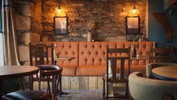 A cozy interior with a stone wall, a tufted orange sofa, wooden chairs, and tables.