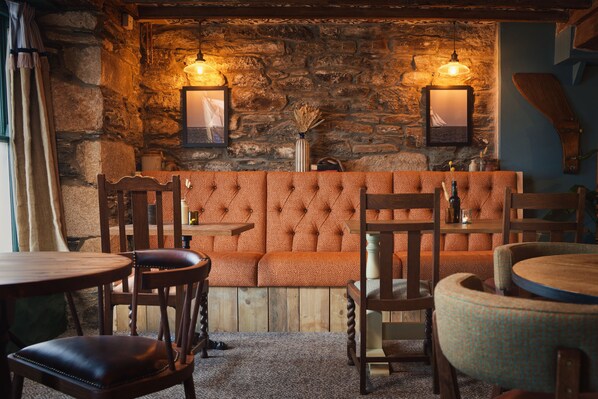A cozy interior with a stone wall, a tufted orange sofa, wooden chairs, and tables. - Wellington Hotel (Boscastle)