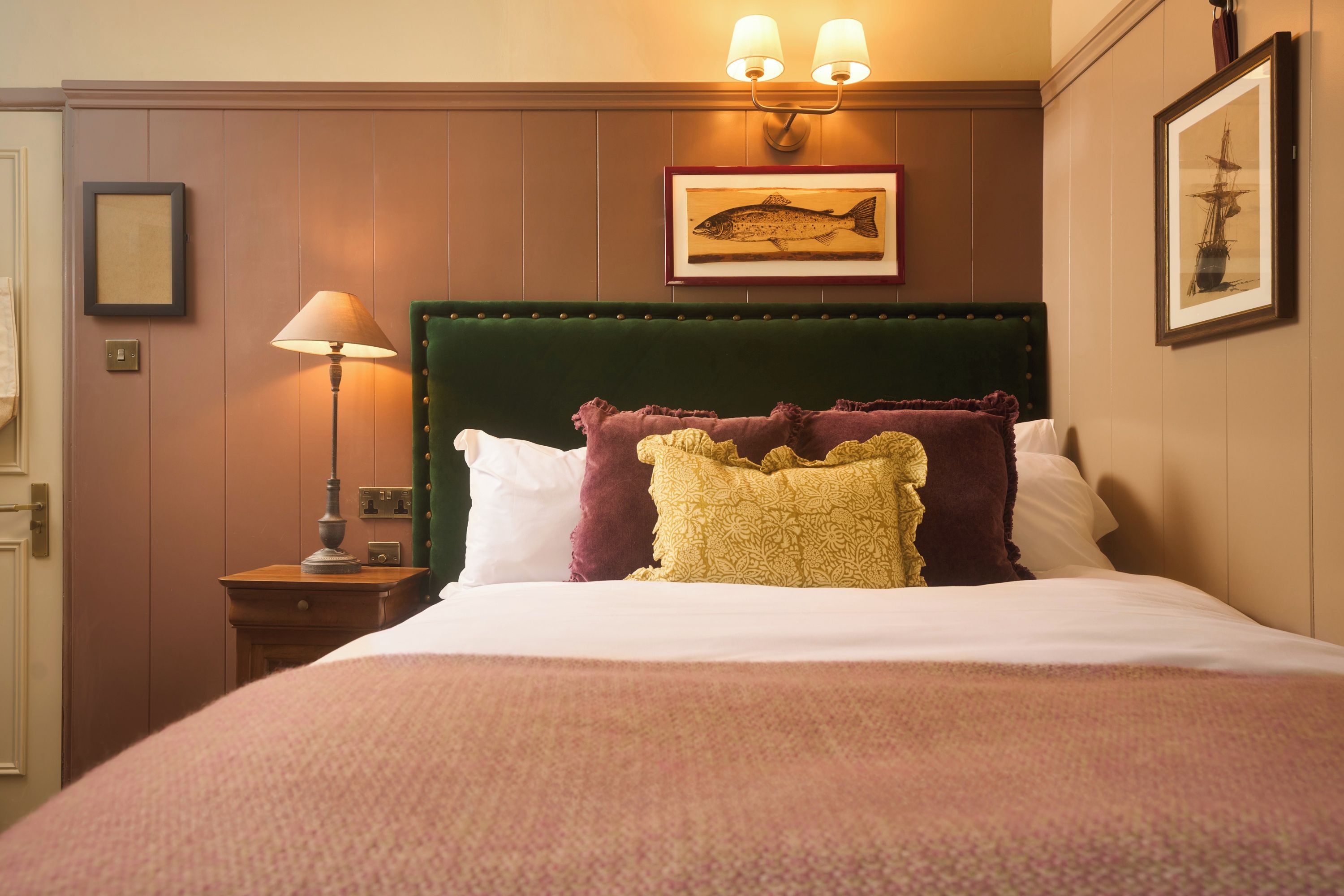 Une chambre d’hôtel avec un grand lit, une tête de lit recouverte d’un tissu vert, une lampe fixée au mur et des œuvres d’art encadrées.