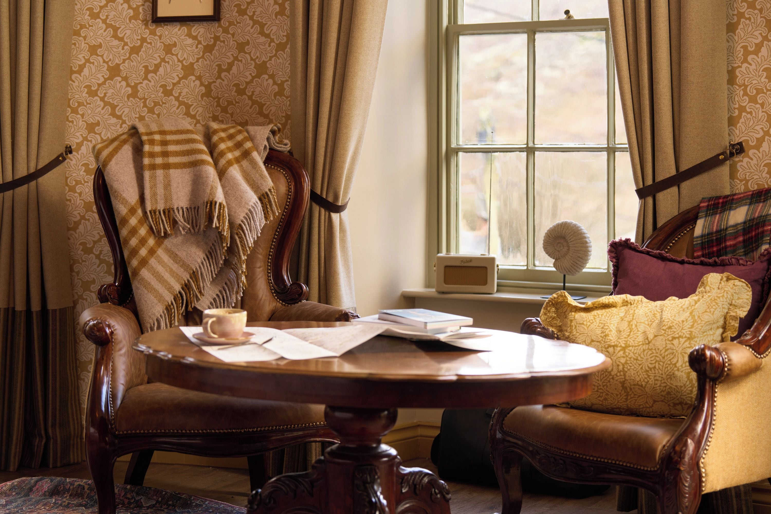 A room with a round wooden table, two chairs, a sofa, and a window with curtains.