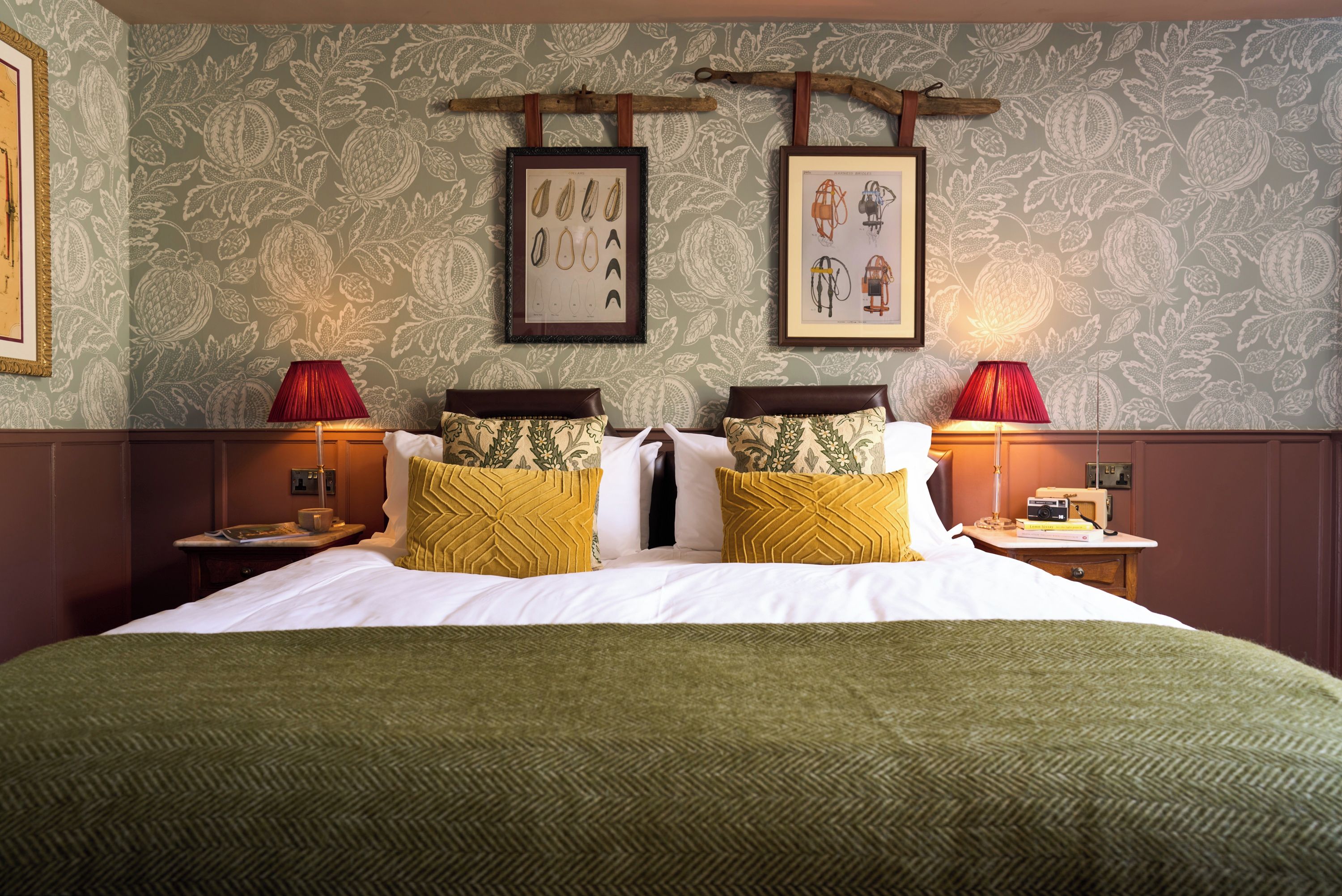 Habitación de hotel con una cama grande, dos mesitas de noche, una lámpara roja y una pared con ilustraciones botánicas enmarcadas.