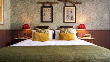Habitación de hotel con una cama grande, dos mesitas de noche, una lámpara roja y una pared con ilustraciones botánicas enmarcadas.