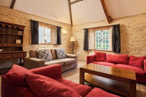 A cozy room with two sofas, a wooden coffee table, and a lamp. There are windows with curtains, a wooden shelf, and a stone wall.