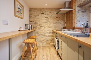 A compact kitchen with a stone wall, wooden stools, a stainless steel sink, and a kettle on the counter. - Wellington Hotel (Boscastle)