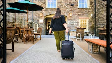 A person walking with a suitcase in a courtyard with stone walls, wooden furniture, and outdoor seating.