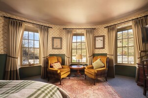 A room with two armchairs, a small table, and a bed. There are two windows with curtains, and the walls have patterned wallpaper.