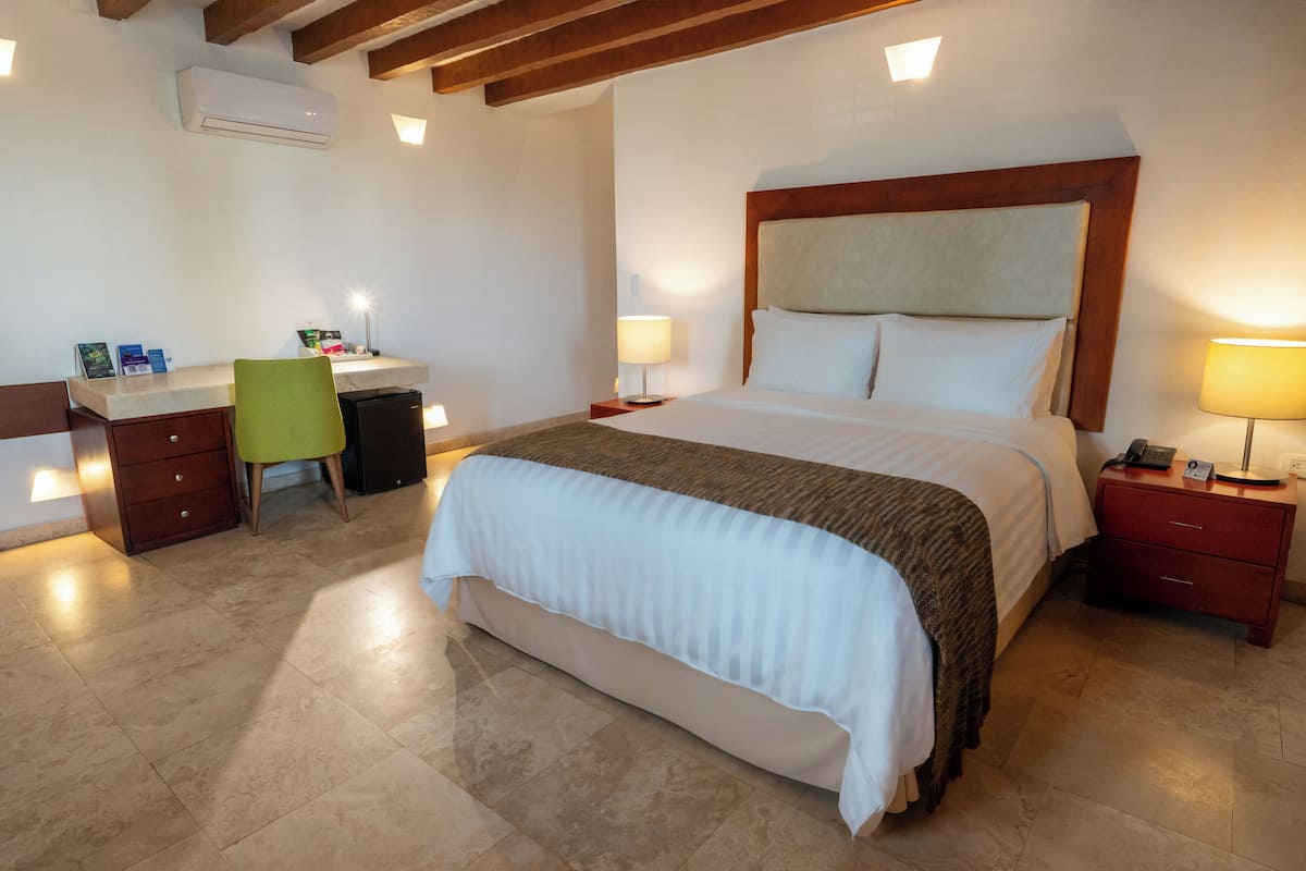 A hotel room with a large bed, a desk with a chair, a bedside table with a lamp, and a red nightstand.