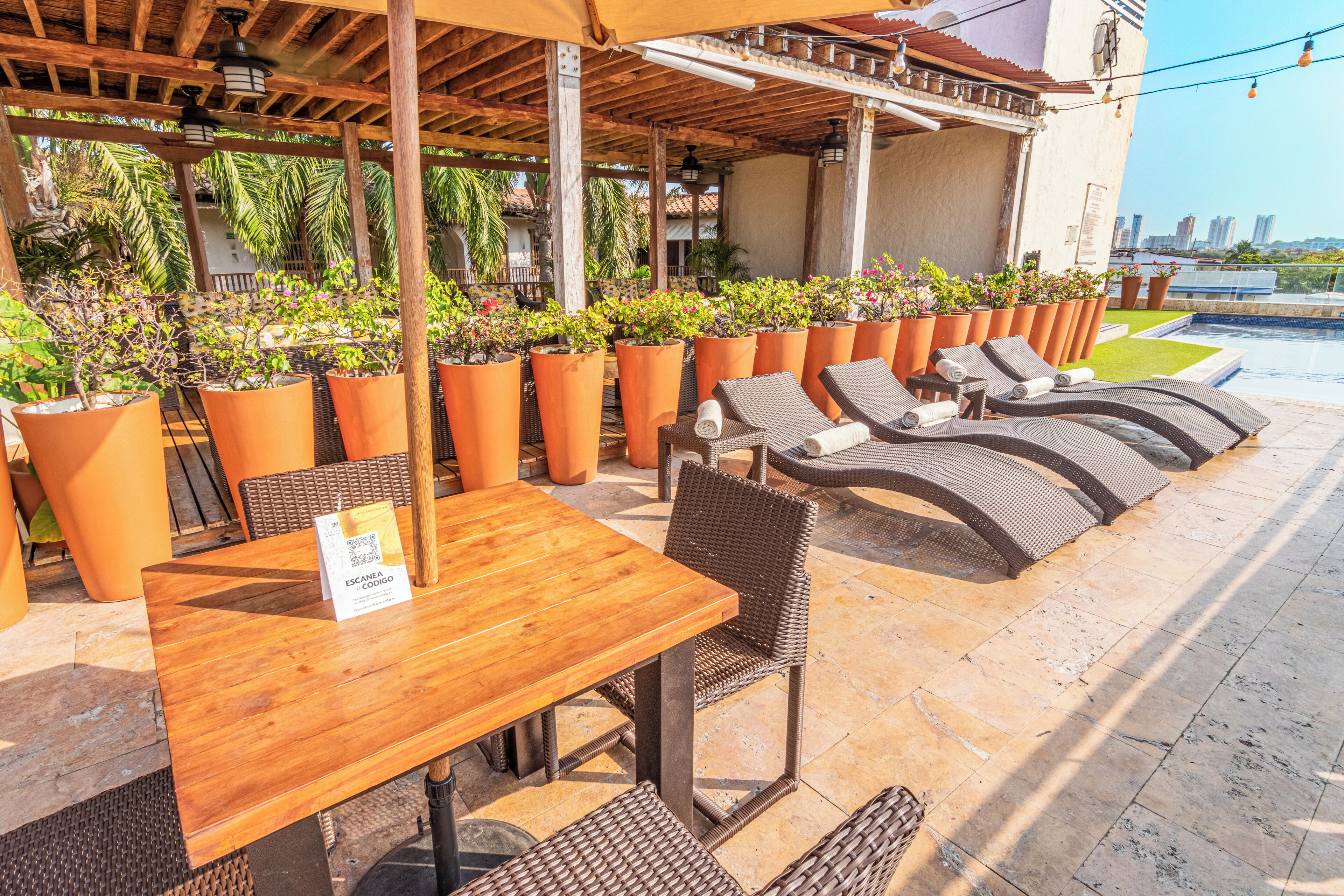 An outdoor seating area with wooden tables and wicker chairs, surrounded by large planters with various plants.