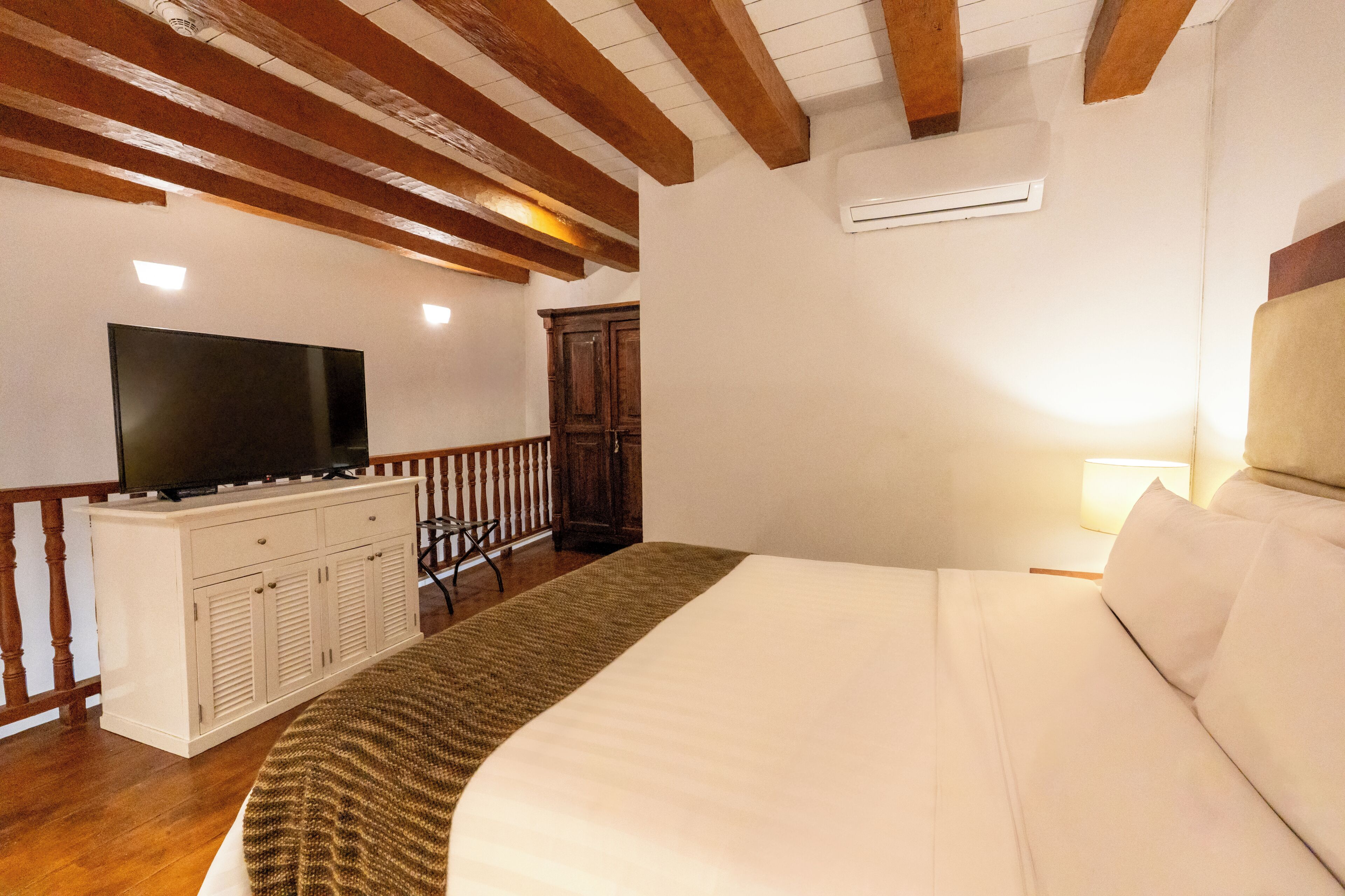 A bedroom with a large bed, a TV on a cabinet, wooden beams on the ceiling, and a nightstand with a lamp.