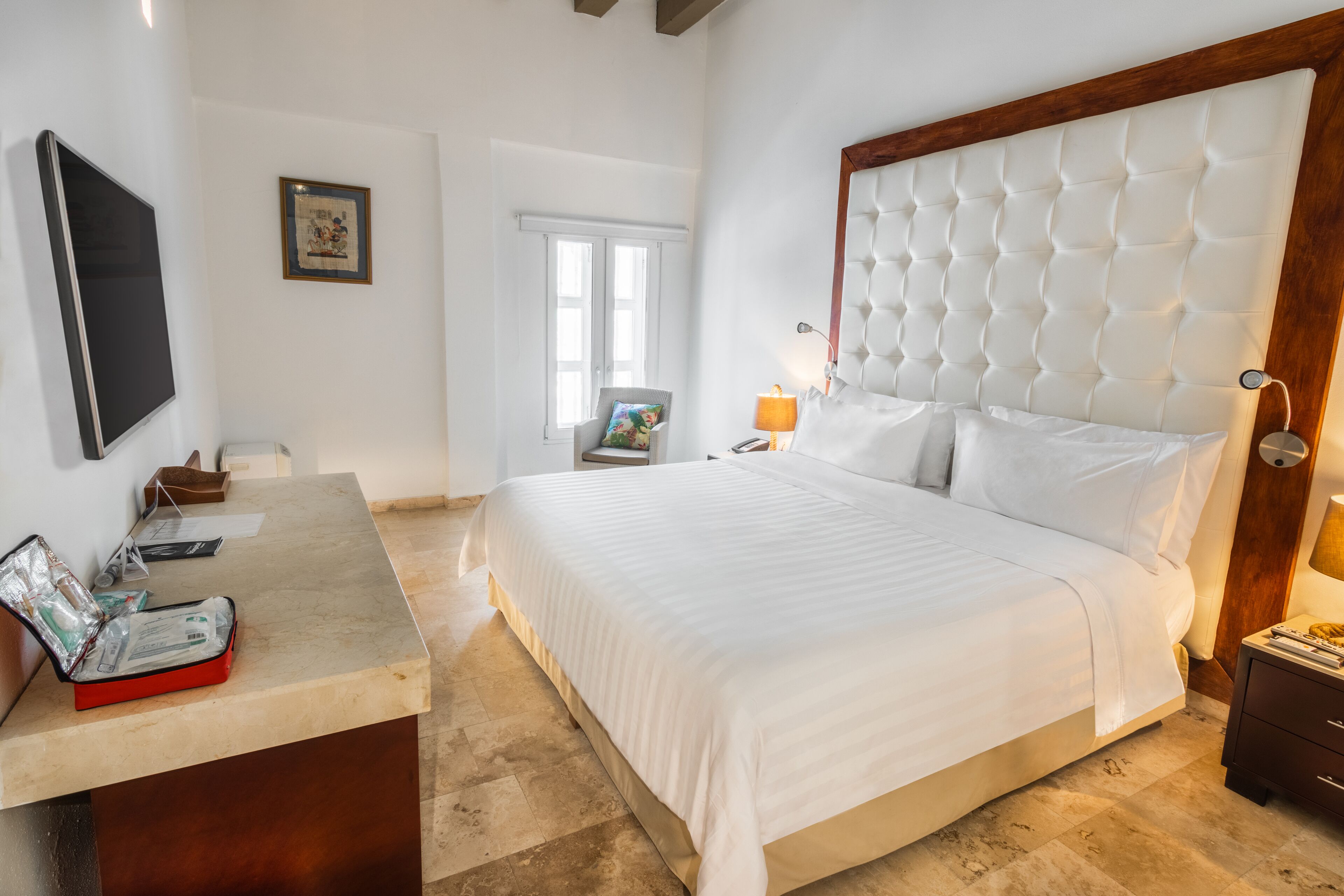 Une chambre d’hôtel avec un grand lit, un bureau avec une mallette rouge, une chaise, une télévision et une fenêtre.