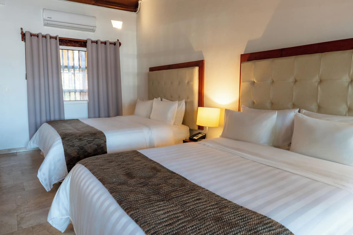 A hotel room with two beds, a window with curtains, a bedside table with a lamp, and an air conditioning unit.